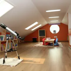 Jasne lampy na poddasze skosy oświetlają pokój z teleskopem, książkami i kącikiem do nauki.