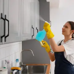 Kobieta w żółtych rękawiczkach spryskuje szafki kuchenne, pokazując, czym czyścić meble kuchenne z okleiny.