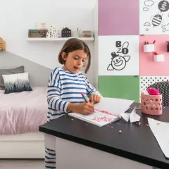 Dziewczynka rysuje w swoim małym pokoju dla dziecka. Na biurku leżą kolorowe kamyczki i papier.