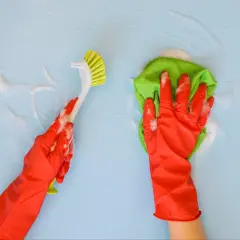 Ręce w czerwonych rękawiczkach czyszczą niebieską ścianę. Jedna ręka trzyma szczotkę, druga zieloną ściereczkę. Idealne do pytania: czym umyć ściany w kuchni.