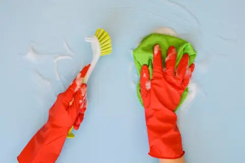 Ręce w czerwonych rękawiczkach czyszczą niebieską ścianę. Jedna ręka trzyma szczotkę, druga zieloną ściereczkę. Idealne do pytania: czym umyć ściany w kuchni.